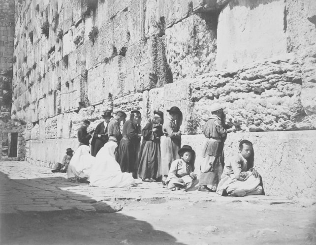 A group of people dressed in traditional garments are gathered against a large stone wall. Several individuals are standing, while others sit or kneel on the ground. The wall shows signs of weathering and has plants growing between the stones. It appears to be a public area with a historical or religious significance.