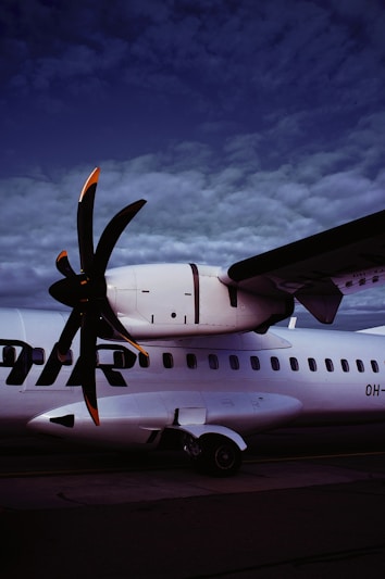A close-up view of an aircraft's turboprop engine and part of the wing against a dramatic sky. The propeller blades have orange tips, and the aircraft body is white with black markings.