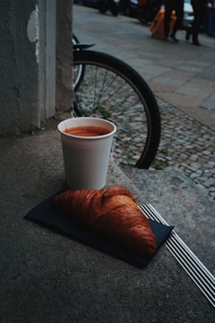 A paper cup filled with coffee is placed on a stone surface beside a freshly baked croissant on a napkin. In the background, a bicycle wheel and a cobblestone sidewalk are visible, with several pedestrians walking past.