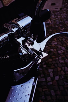 A close-up view of a chrome motorcycle handlebar with a shiny, reflective surface, set against a background of cobblestone pavement.