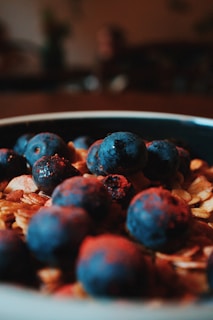 A close-up view of a bowl filled with granola and fresh blueberries. The focus is on the blueberries in the foreground, with their deep blue color contrasting sharply against the light brown of the granola oats.