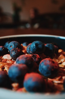 A close-up view of a bowl filled with granola and fresh blueberries. The focus is on the blueberries in the foreground, with their deep blue color contrasting sharply against the light brown of the granola oats.