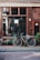A vintage bicycle with a basket is parked on a narrow cobblestone street in front of a shop window. The facade is tiled with dark brown tiles, and a variety of potted plants are lined up against the wall. The glass window reflects the surrounding buildings, street, and a vent in the center. A small number plate with the number 24 is mounted on the wall.