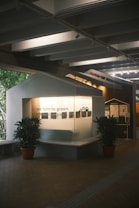 A small exhibit area with informational panels and the phrase 'we turn to green' displayed prominently. The setup is housed under a modern ceiling structure with lines and beams. Two potted plants flank the exhibit, enhancing the natural theme. Ambient light creates a calm and focused atmosphere.