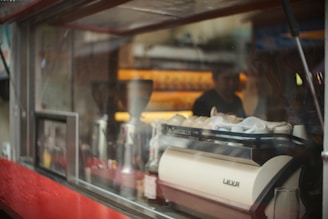 A coffee shop scene with a prominent espresso machine labeled 'Leva' surrounded by various coffee-related items. The background is slightly blurred, showing a person working behind the counter. Reflections appear on the glass panel, adding a layered effect to the image.