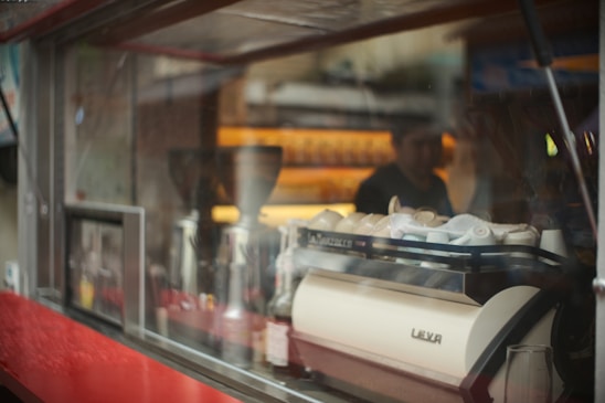 A coffee shop scene with a prominent espresso machine labeled 'Leva' surrounded by various coffee-related items. The background is slightly blurred, showing a person working behind the counter. Reflections appear on the glass panel, adding a layered effect to the image.