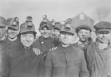 A group of young male miners wearing work jackets and headlamps smile towards the camera. They appear to be taking a break from their work, standing closely together in a friendly and relaxed manner.