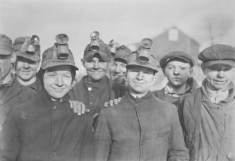 A group of young male miners wearing work jackets and headlamps smile towards the camera. They appear to be taking a break from their work, standing closely together in a friendly and relaxed manner.