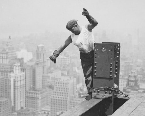 A construction worker balances precariously on a steel beam high above a city skyline. He is wearing a cap, gloves, a white sleeveless shirt, and dark trousers. The background features a dense cluster of tall buildings shrouded in mist or haze, suggesting a high altitude.