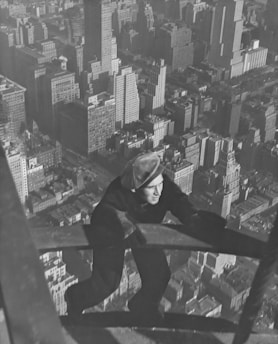 A man is climbing a high structure, possibly a steel beam or ladder, with an expansive view of a city skyline beneath him. The cityscape is filled with tall buildings and skyscrapers, suggesting an urban environment.