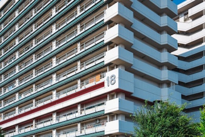 A large, modern apartment building with a series of horizontal lines created by balconies and railings. The structure is predominantly white with accents of green and red. A number 18 is prominently displayed on the side of the building. The angle of the photo emphasizes the geometric patterns in the architecture. There is some greenery visible in the bottom right corner.