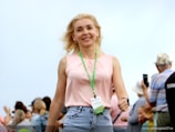 A blonde woman wearing a pink sleeveless top and blue jeans is smiling and walking towards the camera. She has a green lanyard with a name tag around her neck. In the background, several people are visible, some looking at their phones and others conversing, with an out-of-focus setting.