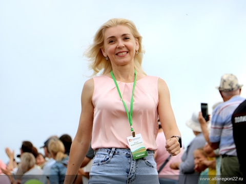 A blonde woman wearing a pink sleeveless top and blue jeans is smiling and walking towards the camera. She has a green lanyard with a name tag around her neck. In the background, several people are visible, some looking at their phones and others conversing, with an out-of-focus setting.