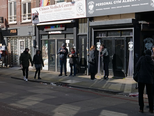 A group of people stand and walk outside a row of storefronts, including a massage center and a personal gym. Some people are using their phones while others are engaged in conversation. A colorful awning adds a pop of color to the street scene, and the clothing style of the pedestrians suggests a cool temperature.