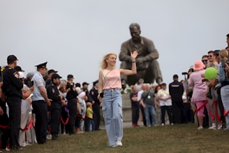 A woman wearing a light pink top and blue jeans is waving her hand while walking on grass. She is surrounded by a crowd of people on both sides, including police officers in uniform, some holding red ropes. Behind her stands a large statue of a sitting figure with crossed arms.