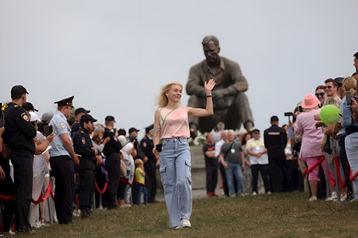 A woman wearing a light pink top and blue jeans is waving her hand while walking on grass. She is surrounded by a crowd of people on both sides, including police officers in uniform, some holding red ropes. Behind her stands a large statue of a sitting figure with crossed arms.