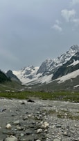 A serene mountainous landscape featuring snow-capped peaks under a clear sky. A river flows through a green valley with rocky edges, surrounded by rugged terrain and patches of grass.