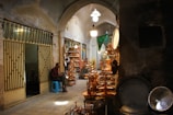 A narrow, dimly lit marketplace or bazaar with arched ceilings. Various copper items are displayed for sale, including trays, teapots, and plates. There is a man in a checkered shirt sitting on a blue stool near the entrance of a shop with striped metal gates. Other shops line the alleyway, filled with more copperware and traditional items. The overall setting appears rustic and historic.