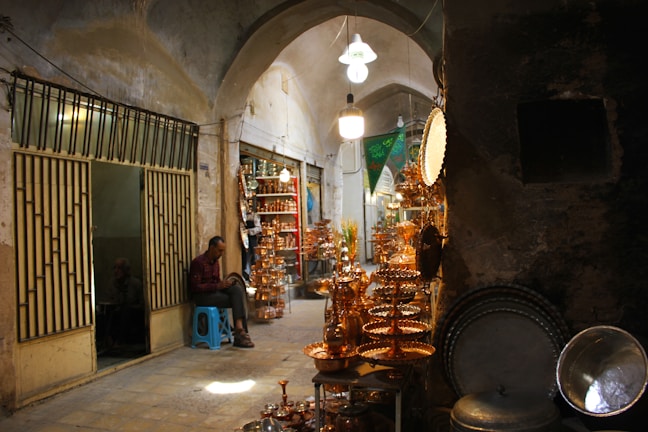 A narrow, dimly lit marketplace or bazaar with arched ceilings. Various copper items are displayed for sale, including trays, teapots, and plates. There is a man in a checkered shirt sitting on a blue stool near the entrance of a shop with striped metal gates. Other shops line the alleyway, filled with more copperware and traditional items. The overall setting appears rustic and historic.