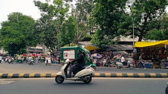 A bustling street scene with trees lining the area. There are multiple vehicles, including motorcycles and auto-rickshaws. A green and yellow canopy covers a section that appears to be a market stall displaying bags and other goods. People are visible moving around on the street and browsing the stalls. The scene is lively and vibrant.