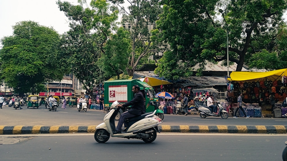 A bustling street scene with trees lining the area. There are multiple vehicles, including motorcycles and auto-rickshaws. A green and yellow canopy covers a section that appears to be a market stall displaying bags and other goods. People are visible moving around on the street and browsing the stalls. The scene is lively and vibrant.