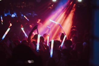 A crowd of people at a lively event holding brightly colored glow sticks. Vibrant lights in red, blue, and purple illuminate the scene, creating an energetic atmosphere.