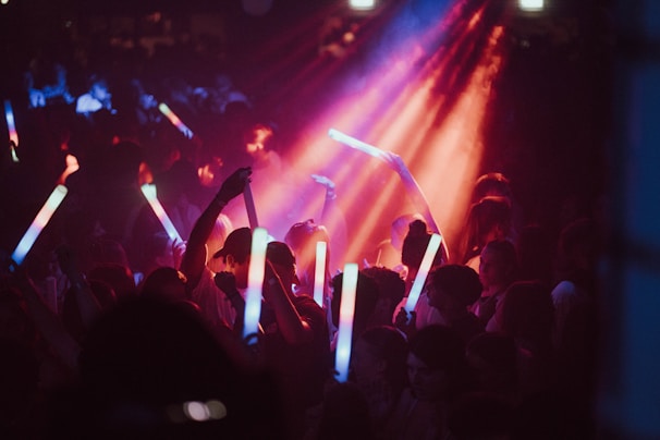 A crowd of people at a lively event holding brightly colored glow sticks. Vibrant lights in red, blue, and purple illuminate the scene, creating an energetic atmosphere.