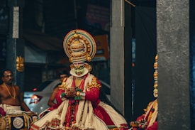 A traditional Indian performance featuring a person in elaborate and colorful costumes and makeup typical of Kathakali dance, with intricate jewelry and a large, ornate headpiece. Other performers are visible, one playing a drum.