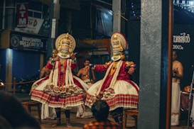 Two performers in traditional Kathakali attire, with elaborate makeup and costumes, engage in a dance performance. The scene includes intricate headgear and vibrant red and white costumes. Men in traditional dhoti observe in the background, and the setting appears to be a cultural or street event with shop signs visible.