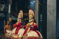 Two performers in elaborate traditional costumes and vibrant face makeup are engaged in a classical dance performance. Their costumes feature intricate patterns, bold colors, and elaborate headgear. The scene captures a cultural or ceremonial event, with a musician playing an instrument in the background.