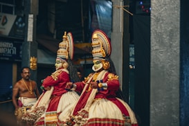 Two performers in elaborate traditional costumes and vibrant face makeup are engaged in a classical dance performance. Their costumes feature intricate patterns, bold colors, and elaborate headgear. The scene captures a cultural or ceremonial event, with a musician playing an instrument in the background.