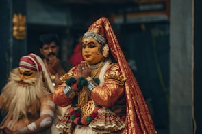 Two people are dressed in traditional costumes, one wearing a vibrant red and gold outfit with intricate patterns, makeup, and jewelry, performing a cultural dance or ritual. The other person has their face painted and wears a cloth with stripes, contributing to the theatrical atmosphere.
