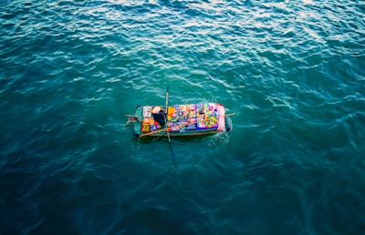 A small boat covered with colorful goods and packages is floating on a vast expanse of calm, dark blue water. The boat appears vibrant against the surrounding water, with various items creating a patchwork of colors atop it.