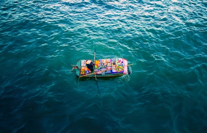 A small boat covered with colorful goods and packages is floating on a vast expanse of calm, dark blue water. The boat appears vibrant against the surrounding water, with various items creating a patchwork of colors atop it.