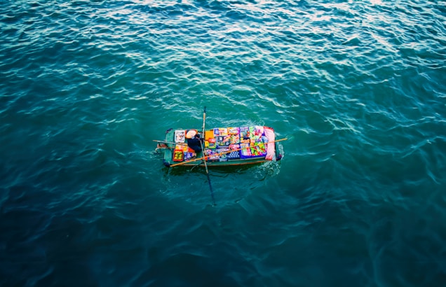 A small boat covered with colorful goods and packages is floating on a vast expanse of calm, dark blue water. The boat appears vibrant against the surrounding water, with various items creating a patchwork of colors atop it.