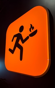A bright orange sign featuring a stylized silhouette of a person running while holding a flaming torch. The sign has rounded corners and is mounted against a dark background.
