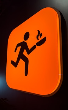 A bright orange sign featuring a stylized silhouette of a person running while holding a flaming torch. The sign has rounded corners and is mounted against a dark background.