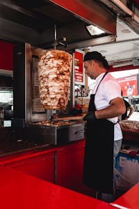 A man dressed in a white shirt and a black apron is slicing a large vertical rotisserie of seasoned meat. The food stand is equipped with red counters and stainless steel appliances. Various kitchen utensils and containers are visible in the background.