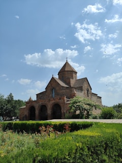 A historic stone church with a cross on top is situated amidst lush greenery under a bright blue sky with scattered clouds. The building features arched doorways and is surrounded by a well-maintained garden with various plants and flowers.