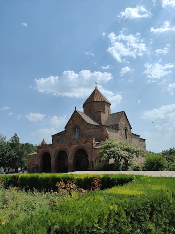 A historic stone church with a cross on top is situated amidst lush greenery under a bright blue sky with scattered clouds. The building features arched doorways and is surrounded by a well-maintained garden with various plants and flowers.