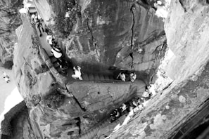 A group of people ascending a narrow, steep stairway carved into a large rock formation with jagged textures. The setting appears to be an outdoor natural environment, possibly a tourist spot, with some vegetation visible along the rock face.