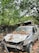 A heavily damaged car with a crumpled front and missing parts is covered in dead leaves, suggesting abandonment. The vehicle is surrounded by dense greenery, indicating it is situated in a forested or overgrown area. The background includes a stone wall and large trees.