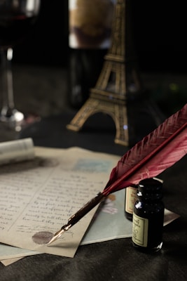 A vintage setting featuring a red quill pen resting on a handwritten letter. Ink bottles and partially visible wine glass and model of the Eiffel Tower are in the background. A warm, dimly lit ambiance evokes a sense of nostalgia and romance.