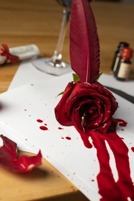 A red rose lies atop a sheet of paper, with red paint or ink pooling around its base, resembling blood. A large red feather is inserted in the rose. In the background, a blurred wine glass and ink bottles are visible, creating an atmosphere of romanticism and mystery.