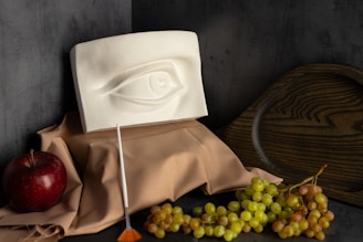 A still life composition featuring a plaster eye sculpture placed on a draped beige cloth. To the left, there is a red apple and a small brush with an orange tip, and to the right, a cluster of green and red grapes. In the background, a wooden, dark plate adds depth to the setup, all against a dark grey textured wall.