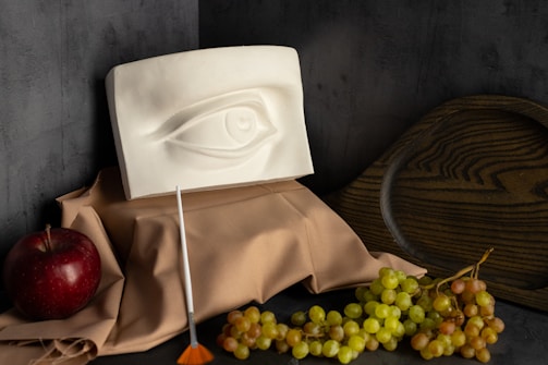 A still life composition featuring a plaster eye sculpture placed on a draped beige cloth. To the left, there is a red apple and a small brush with an orange tip, and to the right, a cluster of green and red grapes. In the background, a wooden, dark plate adds depth to the setup, all against a dark grey textured wall.