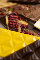 A variety of chocolate bars are adorned with toppings such as dried raspberries and orange peels. The chocolates display geometric patterns and come in different colors including yellow, brown, and dark brown. The toppings add a vibrant and textured appearance to the chocolates.