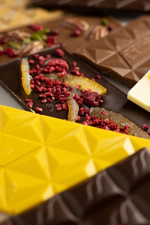 A variety of chocolate bars are adorned with toppings such as dried raspberries and orange peels. The chocolates display geometric patterns and come in different colors including yellow, brown, and dark brown. The toppings add a vibrant and textured appearance to the chocolates.