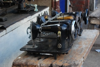 A group of old-fashioned, black Singer sewing machines is placed on a worn wooden table. The machines are lined up in a row, some parts showing rust and wear, indicating their age and usage. The setting appears to be in a rustic or workshop environment with a mix of wooden and metallic surfaces.