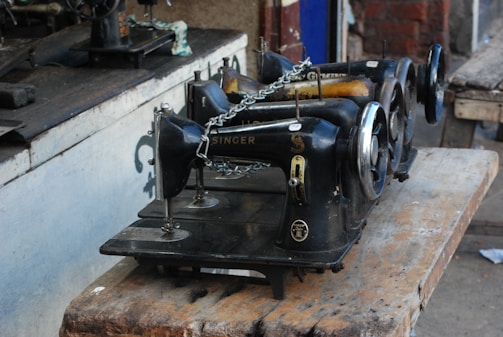 A group of old-fashioned, black Singer sewing machines is placed on a worn wooden table. The machines are lined up in a row, some parts showing rust and wear, indicating their age and usage. The setting appears to be in a rustic or workshop environment with a mix of wooden and metallic surfaces.